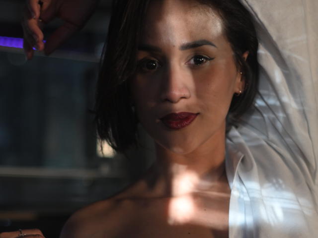 ScarlethFely has medium brown skin, dark brown eyes, and red lipstick, with short brown hair, wearing a white shirt, and a shadowed face with a hand in the top left.