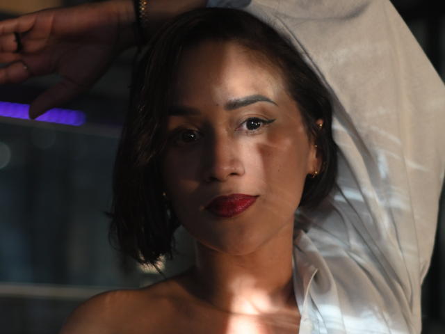 ScarlethFely with dark brown skin, short black hair, red lipstick, and a white shirt, raising her arm, showing a subtle smile, with light and shadow on her face.