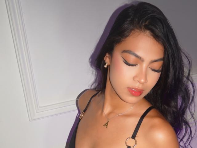 CarolineSantos with closed eyes, dark wavy hair, red lipstick, black strappy top, heart necklace, and earrings, showing off her cute tits and smooth brown skin.