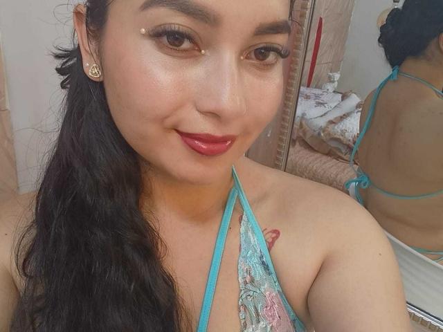 MelaniCoox with long black hair, tan skin, and red lipstick, wearing a blue halter top, showing a small tattoo near her tits, and a nose piercing, smiling at the camera.