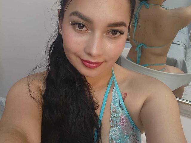 MelaniCoox with long black hair, wearing a blue lace halter top, showing her tits in a mirror selfie, smiling with pink lipstick, and a small tattoo on her right shoulder.