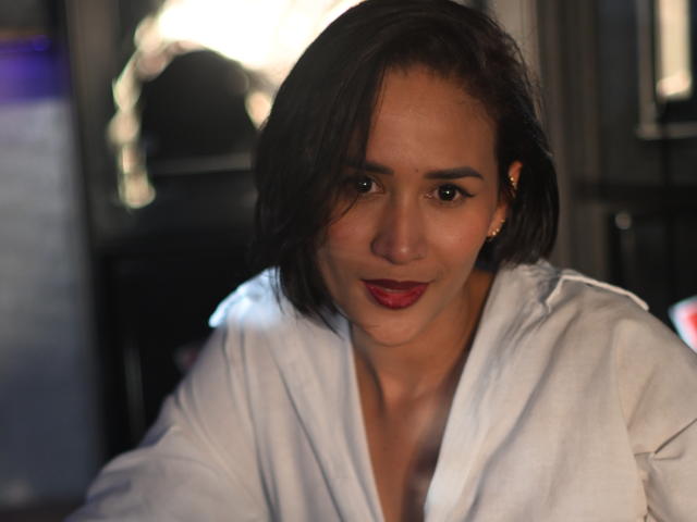 ScarlethFely with short black hair, wearing a white robe, red lipstick, and small gold earrings, has a subtle smile in a dimly lit room.