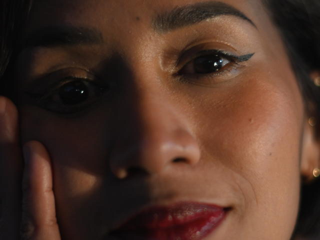 ScarlethFely's face, with dark eyes and black eyeliner, has her hand on her cheek, showing red lipstick and a slight smile. Her skin is tan and textured.