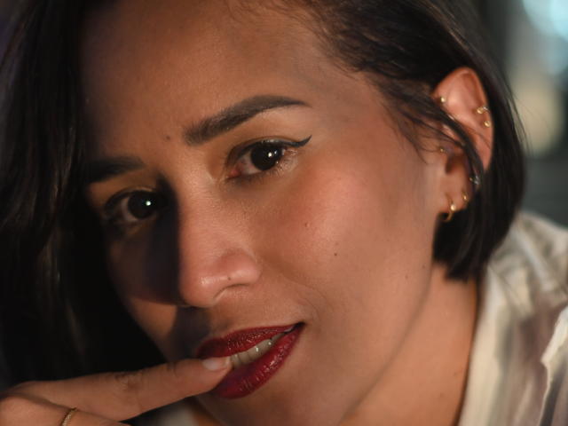 ScarlethFely with dark brown hair, red lipstick, and gold earrings, touching her bottom lip with a finger, showing subtle makeup and a cute, sexy smile.