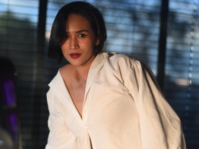 ScarlethFely with short black hair, wearing a loose white shirt, showing her bare shoulders and subtle smile, standing in front of blinds, sunlight casting soft shadows.