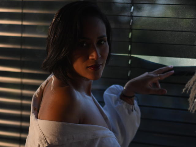ScarlethFely with dark hair, looking over shoulder, wearing off-shoulder white shirt, pointing finger, dim light, shadow on face, subtle smile, ring on finger, dark blinds in background.