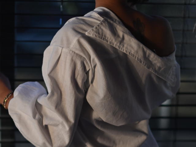 ScarlethFely with dark skin, tattoo on left shoulder, white off-shoulder shirt, sunlight casting shadows, right arm raised, bracelet on wrist, background of horizontal blinds.