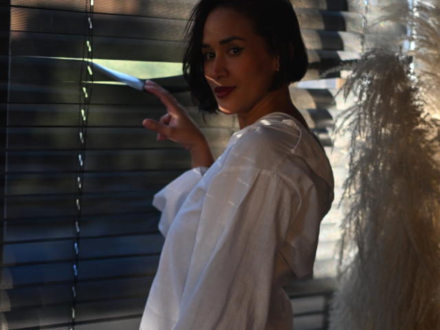 ScarlethFely with short black hair, white shirt, and red lipstick, pointing at blinds, soft light on right, subtle smile, hands near face, shadowed background.