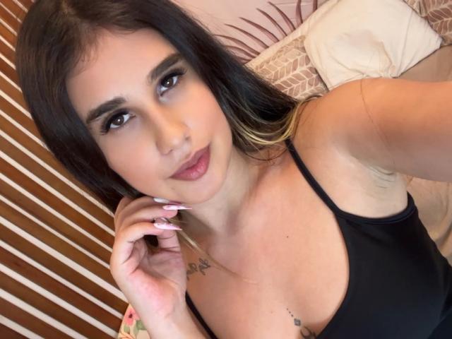 KatieDesirX has long dark hair with blonde tips, wearing a black tank top, showing her tits and tattoos, with pink nails, and a slight smile in a selfie.