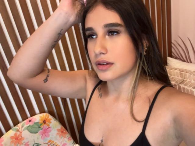 KatieDesirX with brown hair, small tits, tattoos on arm and chest, wearing black top, hand in hair, looking at camera, pink lips, subtle makeup, wooden background.