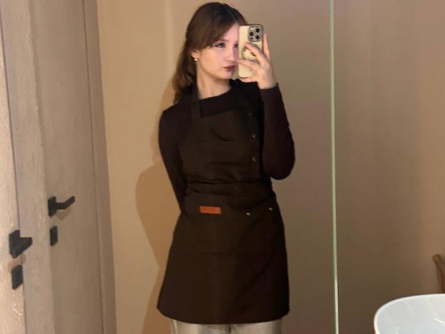 EloisBoeding with brown hair, wearing a black apron over a black long-sleeve shirt, taking a mirror selfie with a phone, showing her ass slightly from the side.
