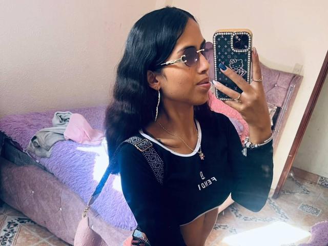 MyaaTyler with dark wavy hair, wearing sunglasses, a black crop top, and a pink bag, taking a selfie, showing her small tits and slim waist.