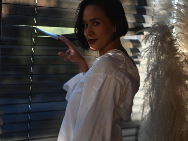 ScarlethFely with short black hair, white loose shirt, pointing at blinds, dark lipstick, soft smile, sunlight highlighting her face, fluffy white decoration on the right.