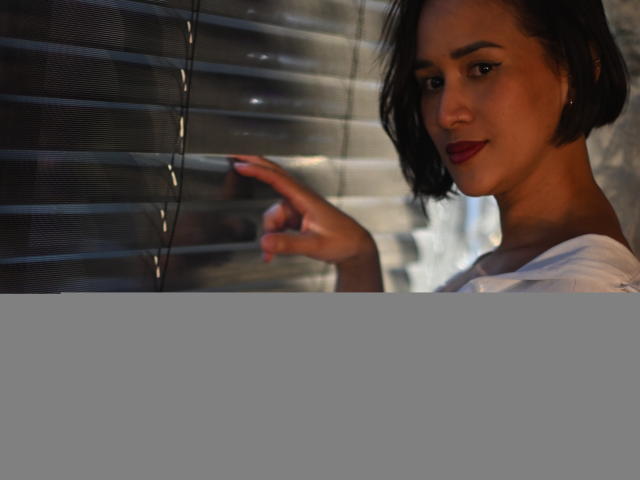 ScarlethFely with short black hair, red lipstick, and white shirt, pointing at metal blinds, her hand near her face, a slight smile on her face, looking sexy.