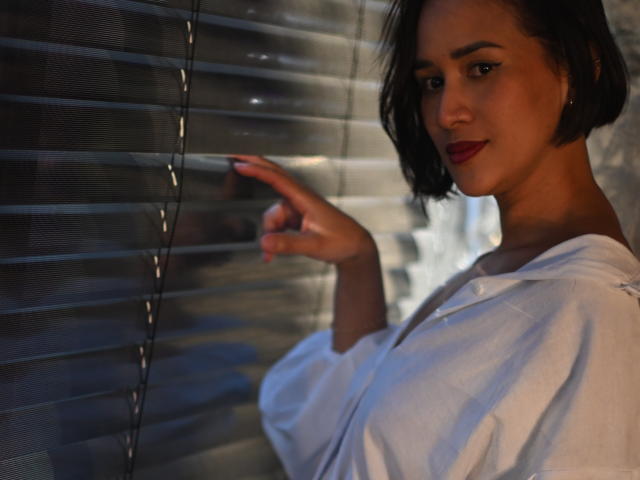 ScarlethFely with short black hair, red lipstick, white shirt, pointing at blinds, dark brown skin, looking slightly to the side, soft lighting, subtle smile, finger pointing gently.