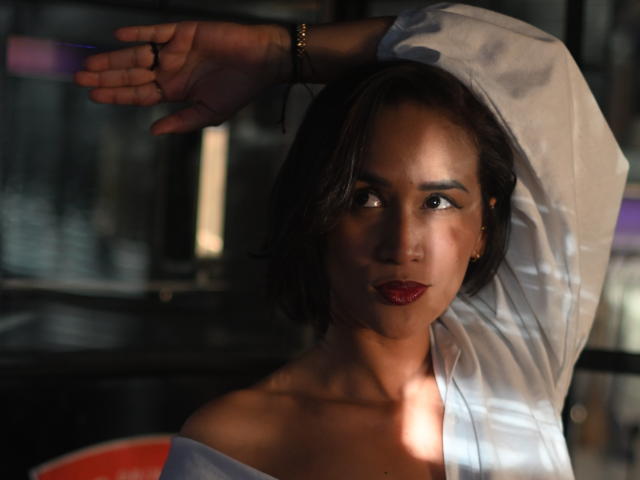 ScarlethFely, avec des cheveux courts et foncés, portant une chemise blanche, bras levé, rouge à lèvres rouge foncé, regardant sur le côté, visage ombragé, bracelet discret au poignet, lumière tamisée, expression sérieuse..