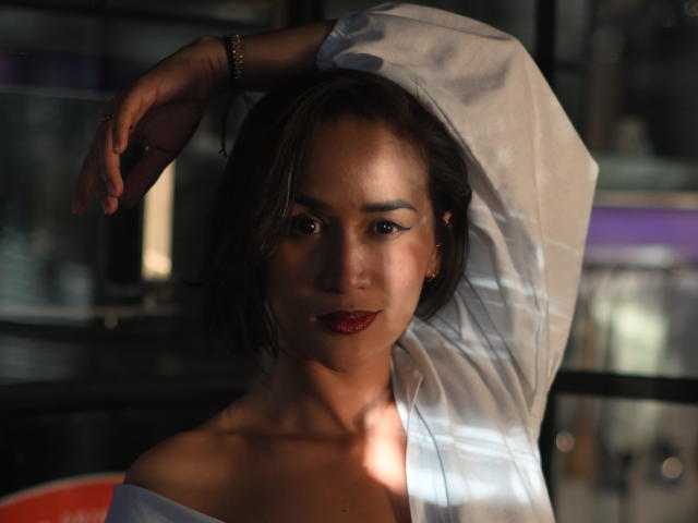 ScarlethFely with dark hair, red lipstick, white shirt, arm raised, shadow on face, subtle smile, light on shoulder, dark background, bracelet on wrist.