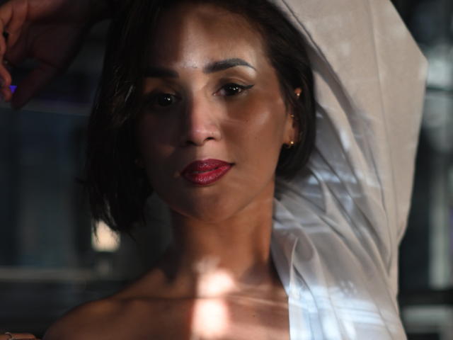 ScarlethFely with dark brown hair, red lipstick, and a white shirt, raising an arm, showing her bare shoulders and a confident, sexy expression.