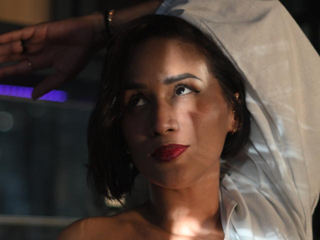 ScarlethFely with dark brown hair, red lipstick, and a white shirt, raising her arm, looking to the side, wearing a gold bracelet, in a dimly lit setting.