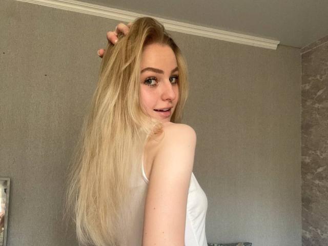 NievesMegee with long blonde hair, white tank top, looking over shoulder, smiling slightly, holding hair up, showing bare shoulder and upper back.