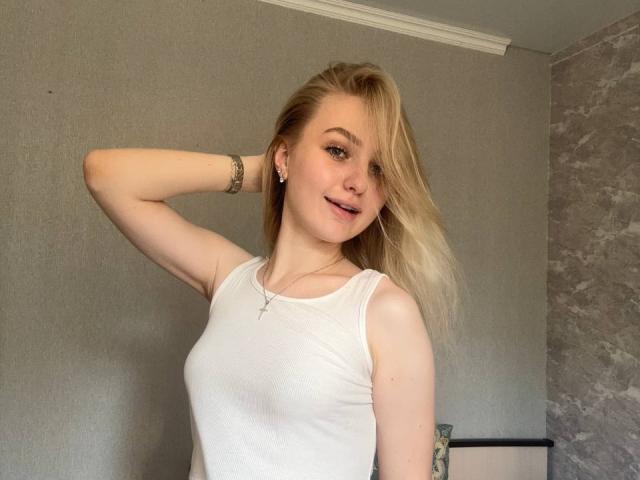 NievesMegee, avec des cheveux blonds, un débardeur blanc, de petites seins, souriante, une main dans les cheveux, un bracelet, des boucles d'oreilles, un collier, une lumière douce, décontractée, sexy, confiante, mignonne..