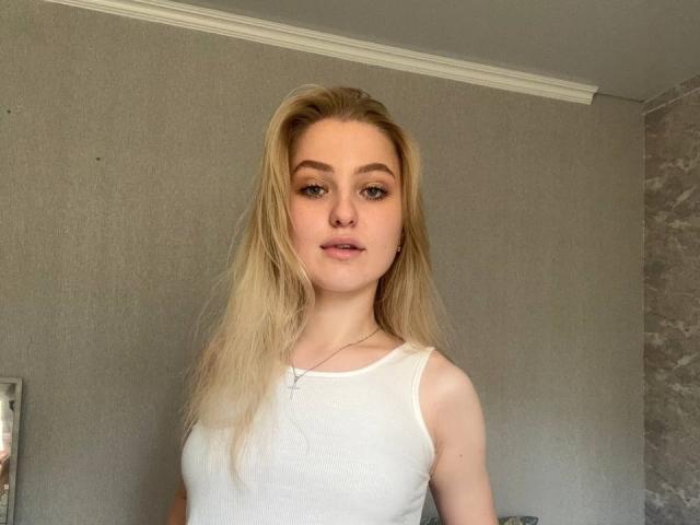 Blonde NievesMegee with fair skin, wearing a white tank top, has small tits and a cross necklace, standing in a gray-walled room, looking at the camera.