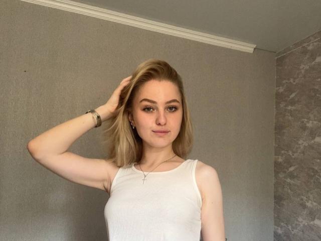 NievesMegee, avec des cheveux blonds, portant un haut blanc sans manches, une main dans les cheveux, souriante, yeux verts, bracelet, collier, mur gris, texture de marbre sur la droite..