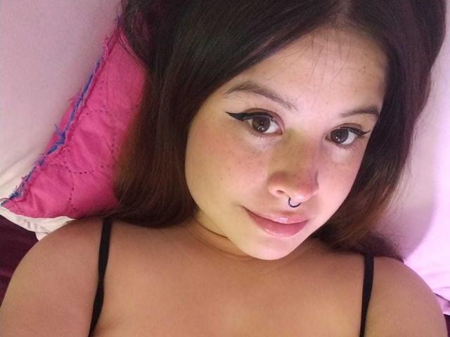 Melani Becker, met bruine ogen, zwarte eyeliner, een neusring en bruin haar, ligt op een roze kussen, draagt zwarte bandjes en kijkt met een lichte glimlach naar de camera..