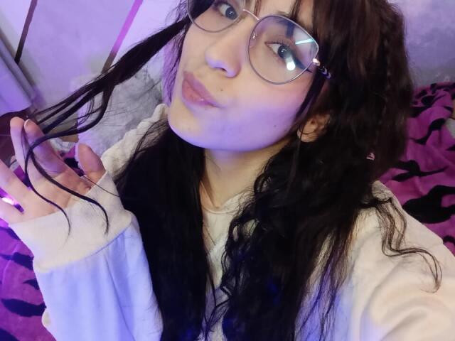 AshlyValdes, avec ses longs cheveux noirs, ses lunettes et ses lèvres pulpeuses, tenant une mèche de cheveux, portant une chemise blanche à manches longues, sur un fond violet et rose..