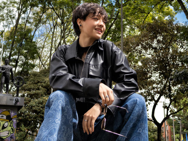 LissyLuxe with curly black hair, wearing a black leather jacket and blue patterned pants, smiling while sitting outdoors, holding glasses in her right hand.