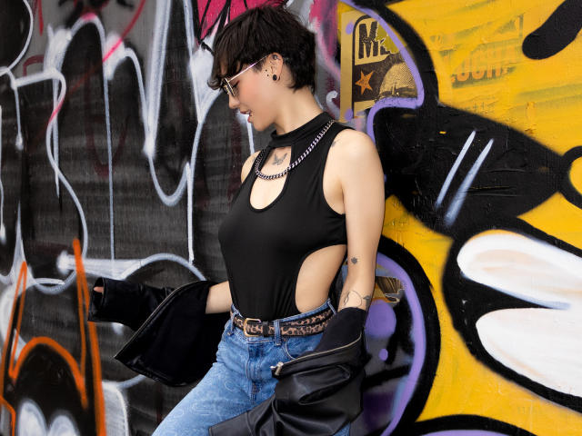 LissyLuxe with short black hair, glasses, black cut-out tank top, belt, jeans, black jacket, and chain necklace, standing in graffiti-covered urban setting. Boobs visible through tank top.