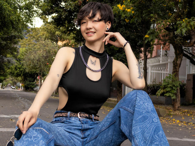 LissyLuxe has short dark hair, a butterfly tattoo on her chest, wears a black sleeveless top, blue patterned jeans, and a chain necklace, sitting on a street.