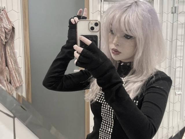 Carmelia Pratten, avec ses cheveux violet pâle, porte une chemise noire à manches longues avec des studs blancs, des gants noirs sans doigts, et prend un selfie devant un miroir, tenant un iPhone doré, dans une salle de bain..