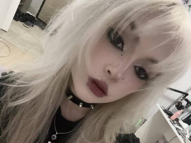 CarmeliaPratten with platinum blonde hair, dark eye makeup, lip studs, chipped black lipstick, and a spiked black collar, looking at the camera with a slightly open mouth.