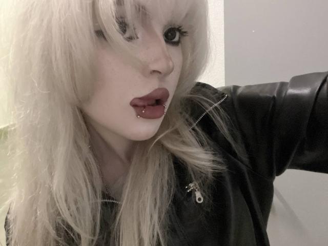 Carmelia Pratten, avec ses cheveux blancs et en bataille, sa veste en cuir noir et ses lèvres rouges maquillées, regarde fixement avec un maquillage sombre, tout en levant son bras. Elle a la peau pâle et porte un anneau au nez..