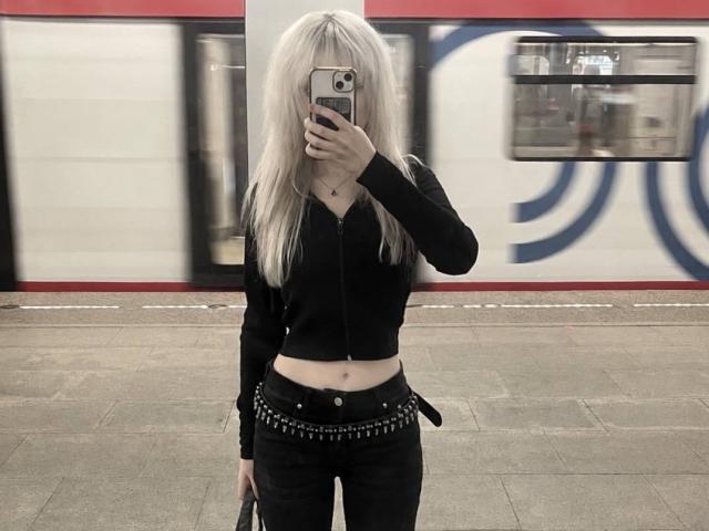 Carmelia Pratten, avec ses cheveux blonds, un haut court noir à fermeture éclair, un pantalon noir, une ceinture avec des pointes, tenant un téléphone, se cachant le visage, debout devant un train flou..