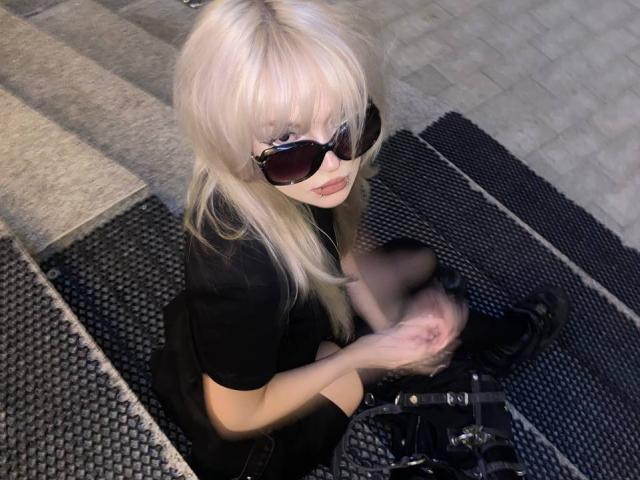 Carmelia Pratten, con cabello rubio largo, usando gafas de sol negras y un atuendo negro, sentada sobre una esterilla negra texturizada, ajustándose las botas negras, mirando hacia arriba con una ligera sonrisa..