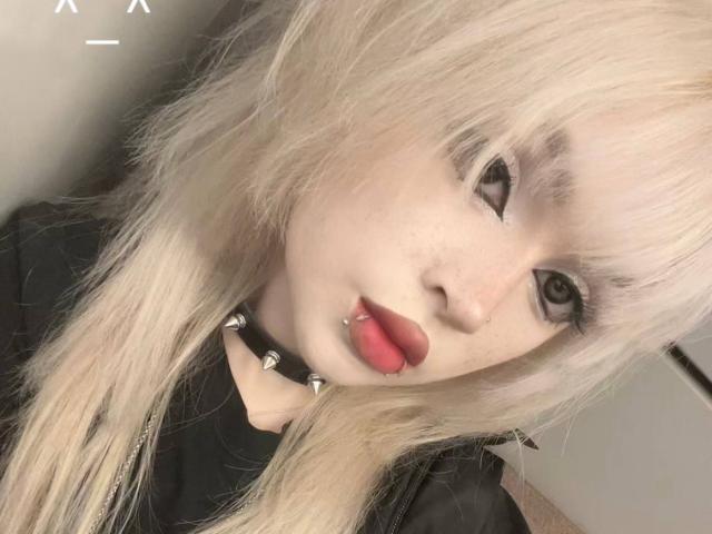 Carmelia Pratten, met lang blond haar, zwarte eyeliner en rode lippenstift, draagt een zwarte choker met metalen studs en een zwart shirt, waarbij een lip-piercing zichtbaar is..
