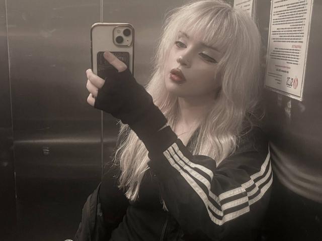CarmeliaPratten with blonde hair, wearing a black Adidas jacket with white stripes, black fingerless gloves, and dark lipstick, taking a selfie in a metallic elevator.