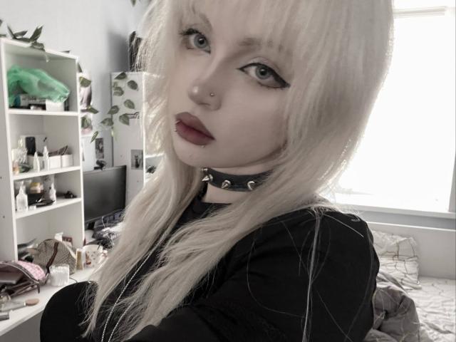 CarmeliaPratten, avec une peau pâle, des cheveux blonds platine, un eye-liner noir, du rouge à lèvres rouge, un anneau au nez, un collier noir, une chemise noire, assise sur un lit, regardant la caméra, avec un fond de chambre..