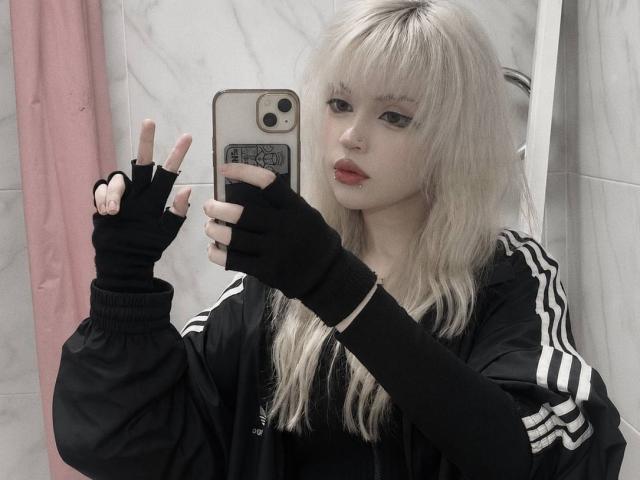 Carmelia Pratten, avec ses cheveux blonds platine, ses gants noirs sans doigts et sa veste Adidas, faisant un signe de la paix tout en prenant un selfie devant un miroir, avec des lèvres rouges et une légère moue..