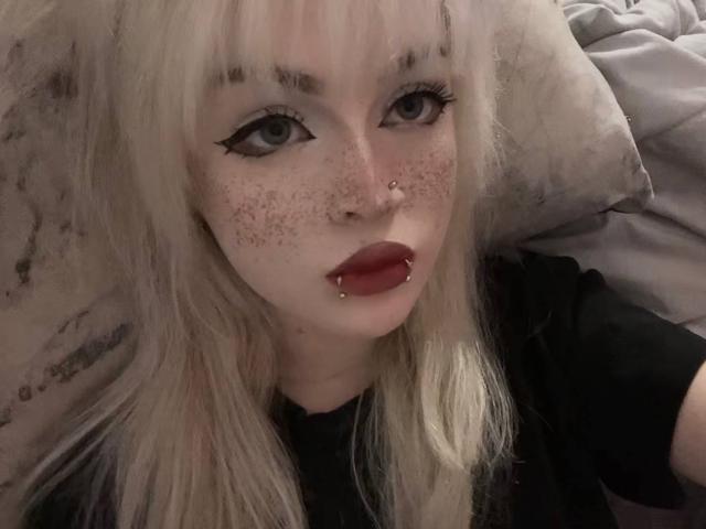 CarmeliaPratten a la peau pâle, les cheveux blonds, du crayon noir pour les yeux, des taches de rousseur, un anneau au nez, des piercings aux lèvres, et du rouge à lèvres rouge, et elle est allongée sur un lit en portant une chemise sombre..