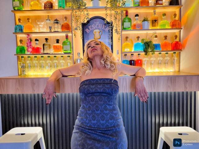 AmelieLux with blonde hair, wearing a blue patterned strapless dress, arms out, standing in front of a colorful liquor shelf, looking up with a slightly annoyed expression.
