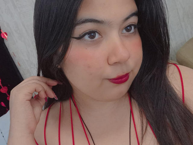 SultanaLm a de longs cheveux noirs, du rouge à lèvres et un trait d'eye-liner noir épais. Elle porte un haut rouge à encolure dégagée, avec une main près de son visage, affichant un sourire subtil..