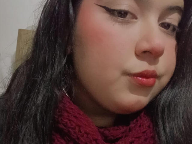 SultanaLm a les lèvres rouges, des ailes de khôl noir, des joues rosées et des cheveux noirs et lisses, elle porte une écharpe rouge en tricot, avec un léger sourire et un regard confiant..