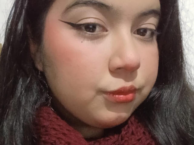 Le visage de SultanaLm, avec ses cheveux foncés, son rouge à lèvres rouge, son eye-liner noir épais, une légère moue, un foulard en tricot rouge, et une petite tache de rousseur sur sa joue gauche..