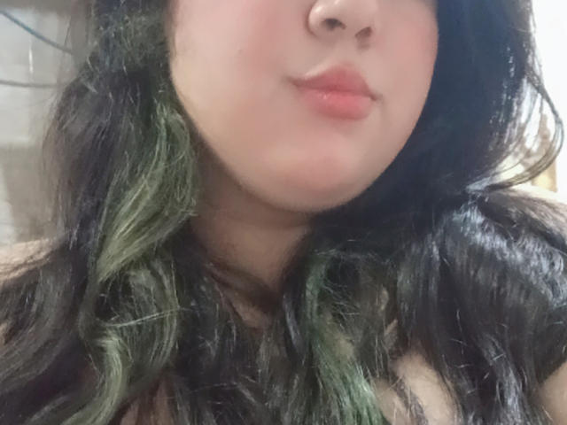 SultanaLm tem pele clara, lábios rosados e volumosos, e cabelo ondulado escuro com mechas verdes, exibindo um leve sorriso. Seu nariz é pequeno e delicado..