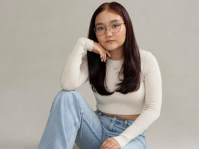 LavonKolsrud, avec des lunettes, de longs cheveux bruns, un crop top blanc à rayures, un jean bleu taille haute, le menton posé sur la main, assise, avec une expression sérieuse, petite poitrine, mignonne, peau claire..