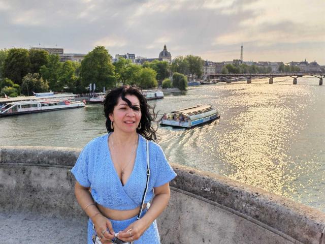 EtoileLatin met een gemiddelde borstmaat, draagt een blauw geruit crop top en een bijpassende rok, en glimlacht naar de camera. Ze heeft zwart, golvend haar en staat bij een rivier met boten..