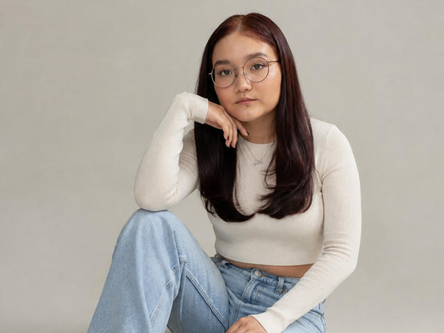 LavonKolsrud, avec des lunettes, un débardeur blanc et un jean bleu, est assise, une jambe levée, la main sur le menton, avec une expression sérieuse. Elle a de longs cheveux bruns foncés..